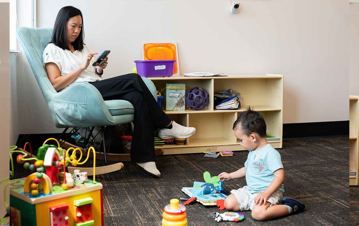 A woman in a white shirt and black pants sits in a green upholstered rocking chair, scrolling on a cell phone, while her toddler sits on the floor playing with toys