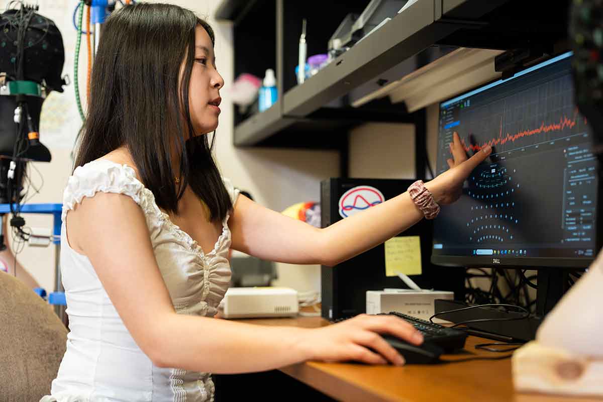 A young woman in a short-sleeved white top sits at a desk in front of a computer screen with scientific data displayed