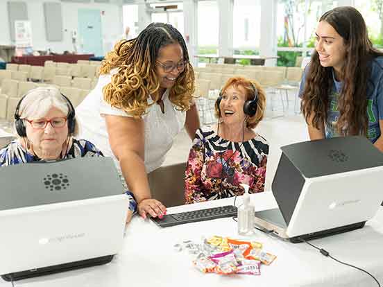 Two older women are seated at computer screens while a professor in a white dress and student in a blue FGCU shirt look on