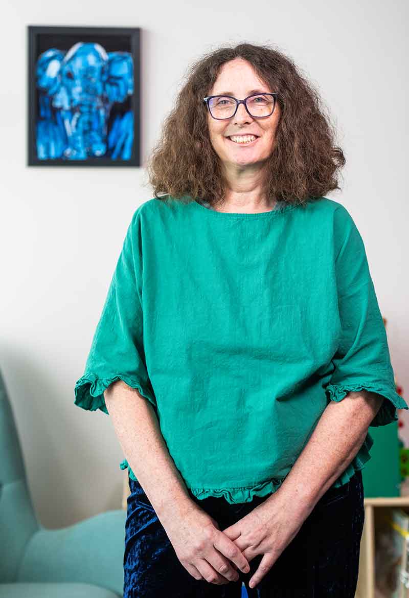 A brunette woman wearing glasses in a green, 3/4-sleeve top and black pants, standing in front of a green upholstered rocking chair