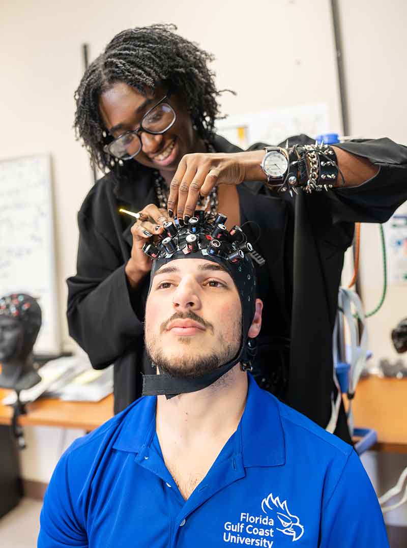 A young woman with glasses in a black shirt fits a skull cap with electrodes and chin strap on a young man in a blue polo shirt