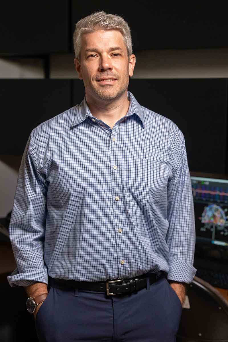A silver-haired man in a light blue button-down shirt and dark blue pants with his hands in his pockets