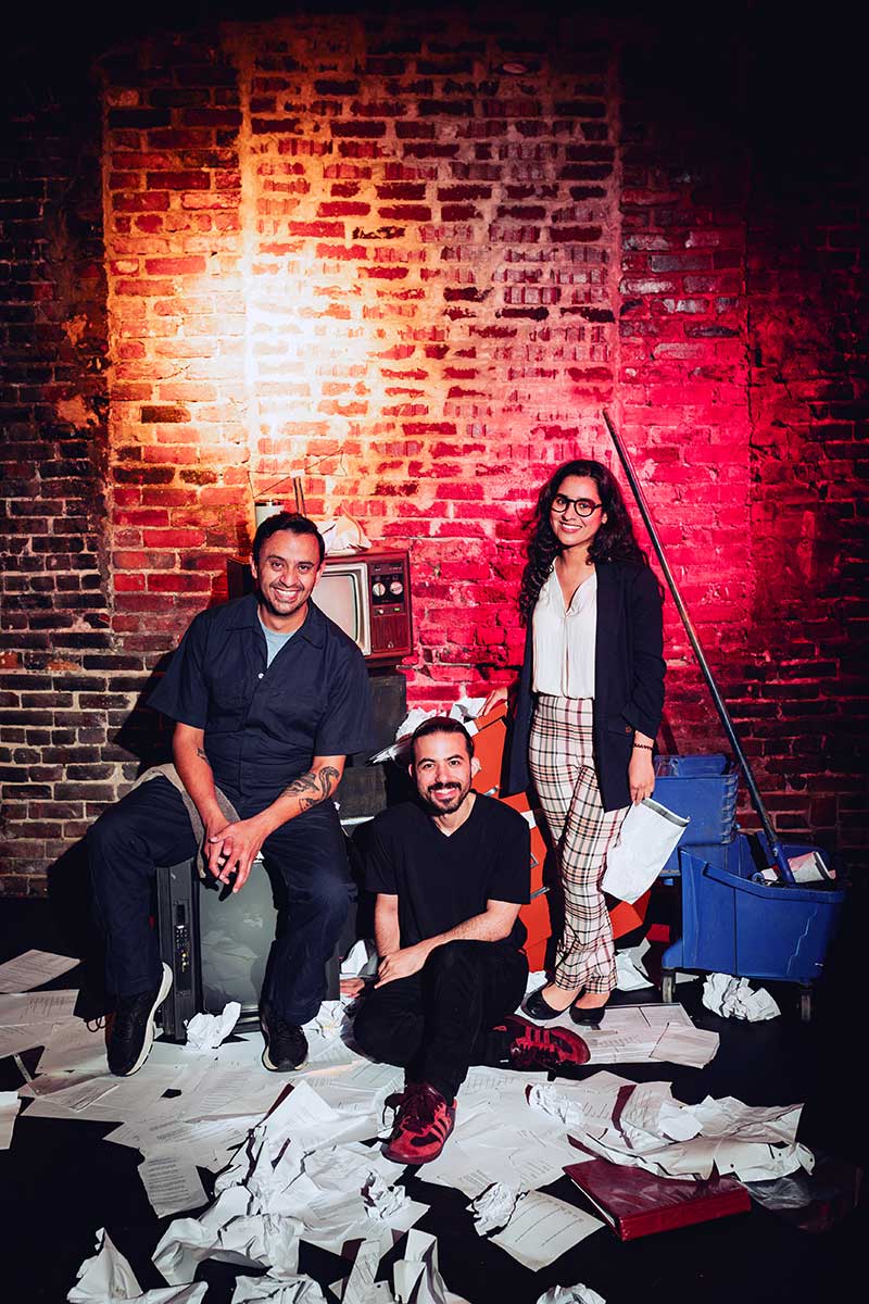 Theater team posing in messy rehearsal room with brick wall and papers