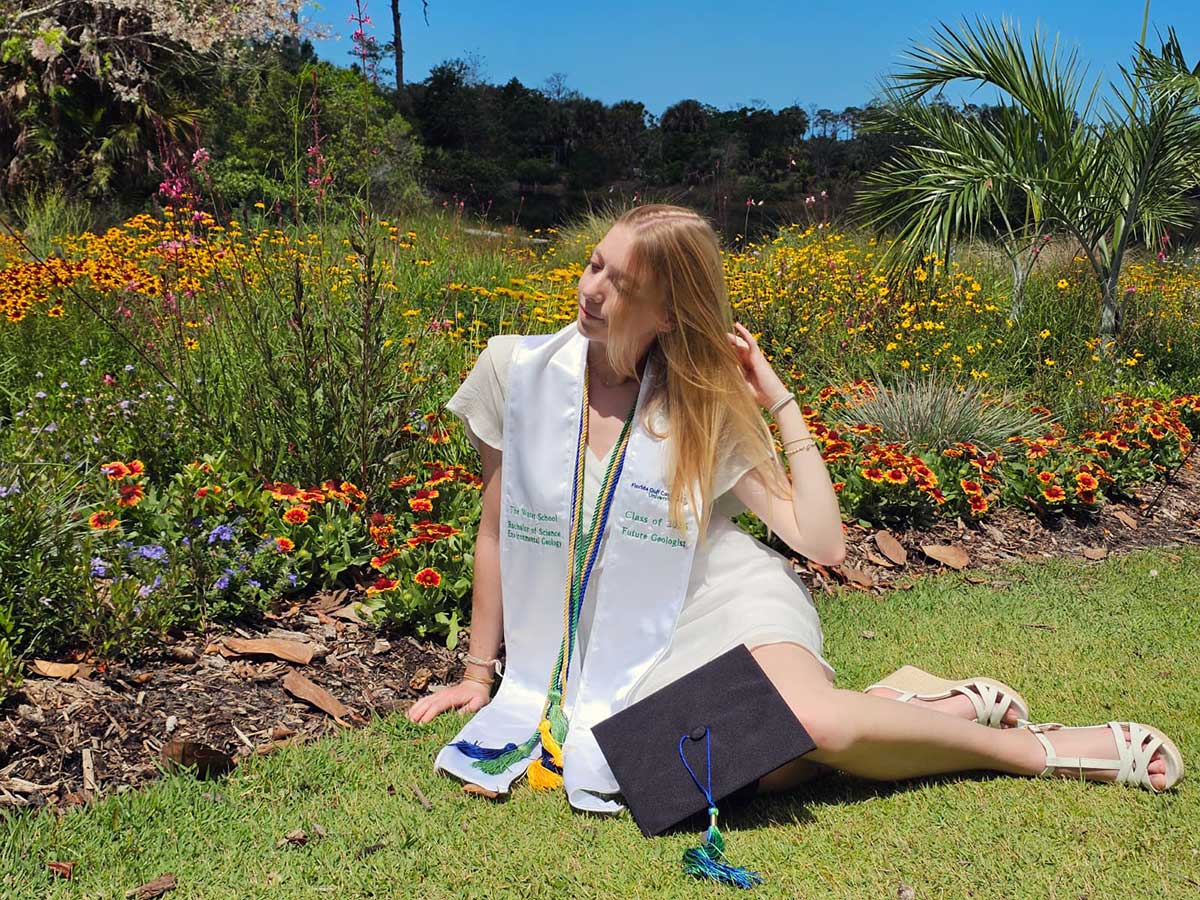 Graduate sitting in flower garden with cap, stole, and diploma outdoors
