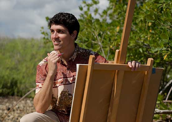 Person sits barefoot on rocky shore with easel and paintbrush, surrounded by greenery.