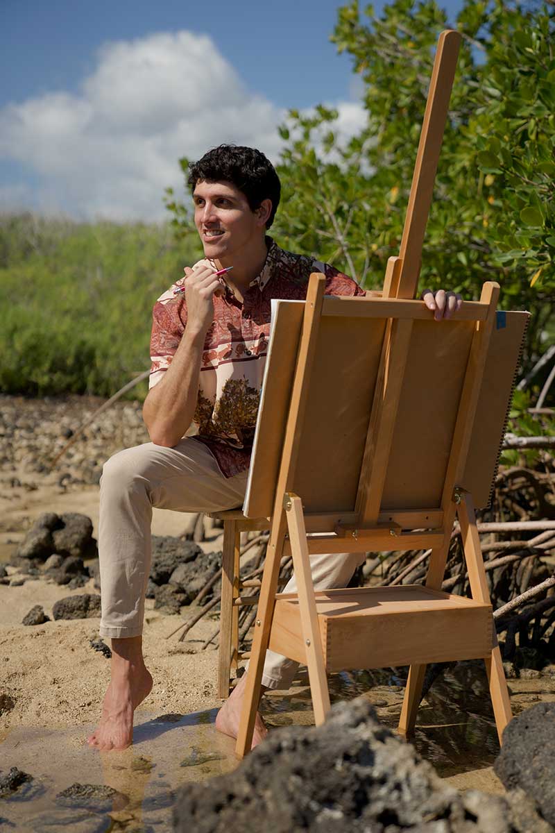 Person sits barefoot on rocky shore with easel and paintbrush, surrounded by greenery.