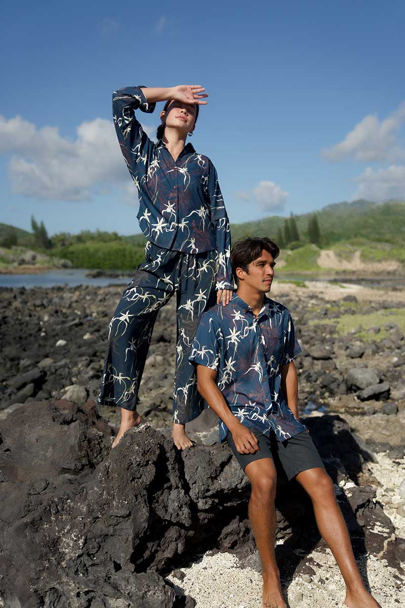 Two people in matching outfits stand and sit on dark rocks by a shoreline under a blue sky.
