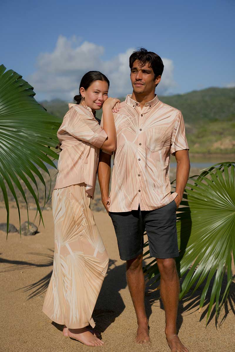 Two adults stand barefoot on a sandy beach with palm leaves, wearing matching outfits.