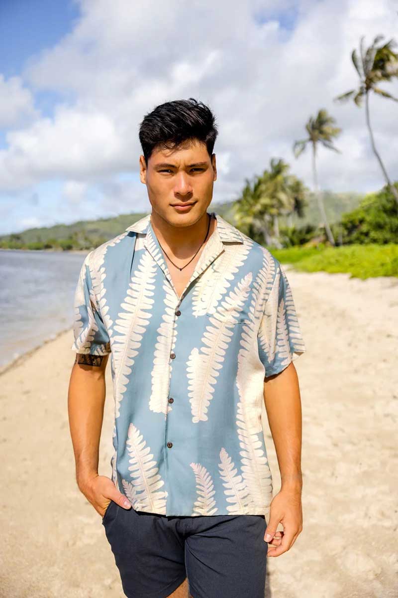 Person stands on sandy beach with palm trees, ocean, wearing a patterned short-sleeve shirt.
