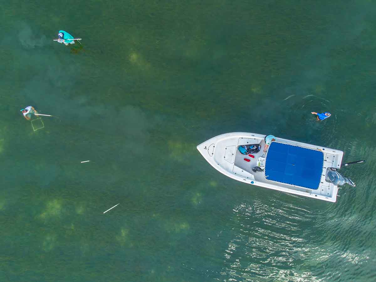 An overhead shot of a boat in a large body of water, with two people in the water and white pipes sticking straight up nearby