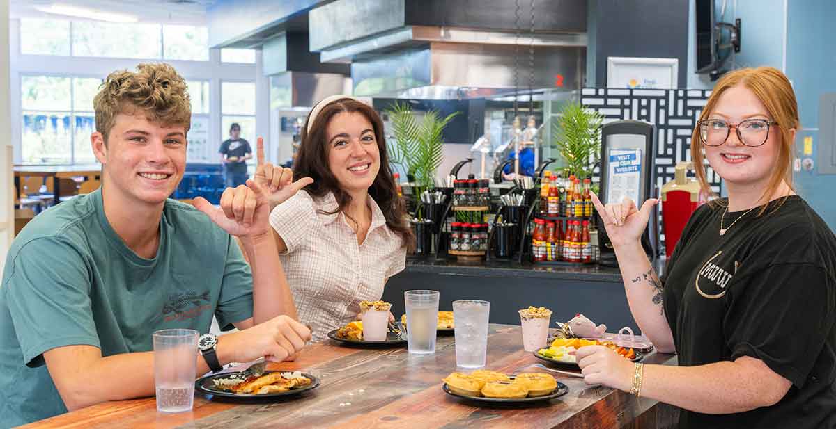 A young man in a green t-shirt, a young brunette in a white, short-sleeved blouse and a young, redheaded woman in a black t-shirt and glasses sit together at a table eating plates of food