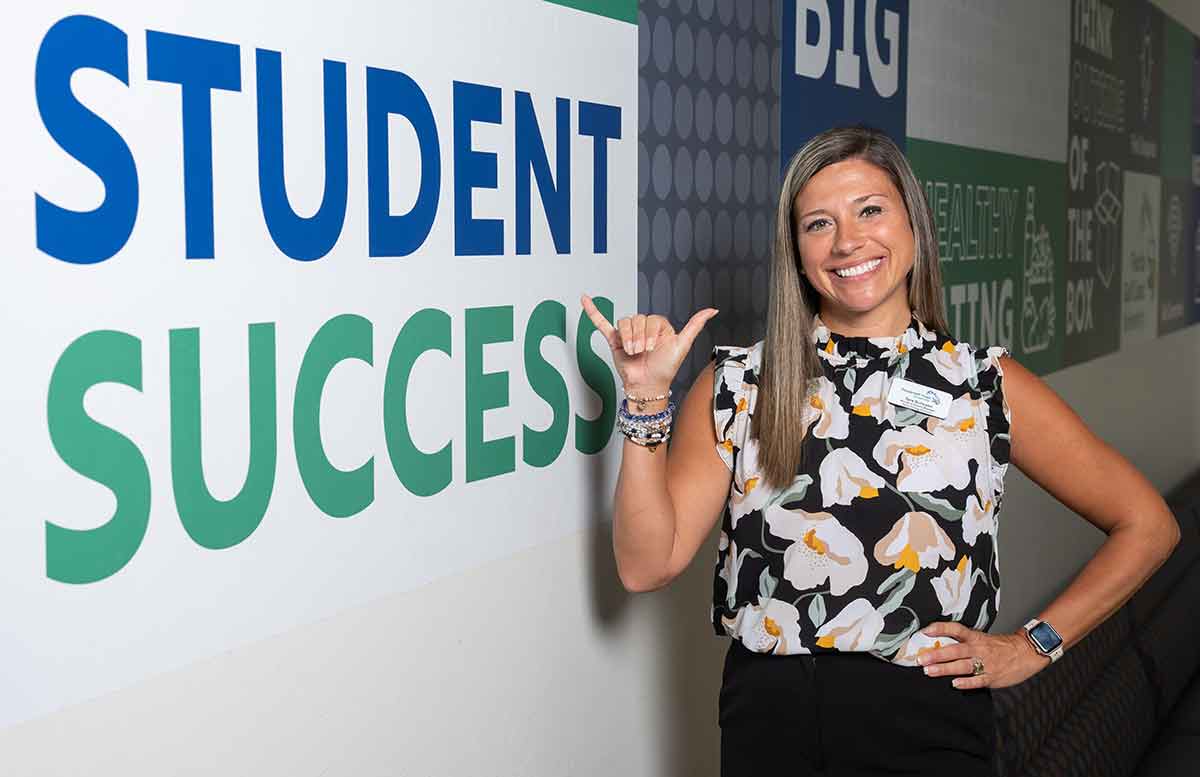 A woman stands, making the FGCU Wings Up gesture with her right hand, in front of a sign that reads Student Success