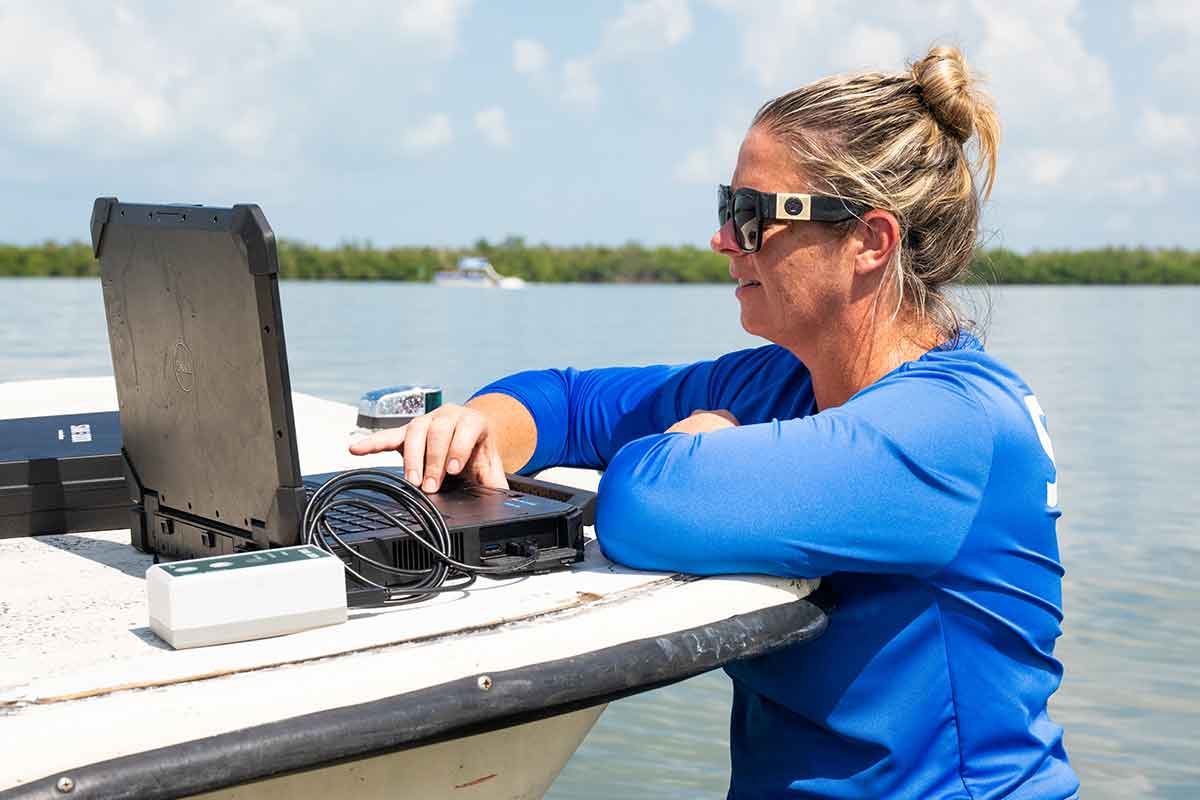 A woman in sunglasses and a blue, long-sleeved shirt, works at a laptop on a boat while she stands off the boat in a body of water, with mangroves and a blue sky in the background