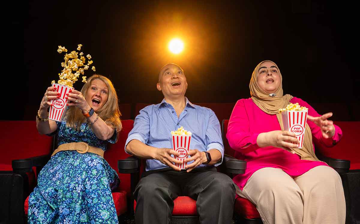 Three people sit in cinema chairs, holding popcorn containers, as if watching a movie, with surprised looks on their faces