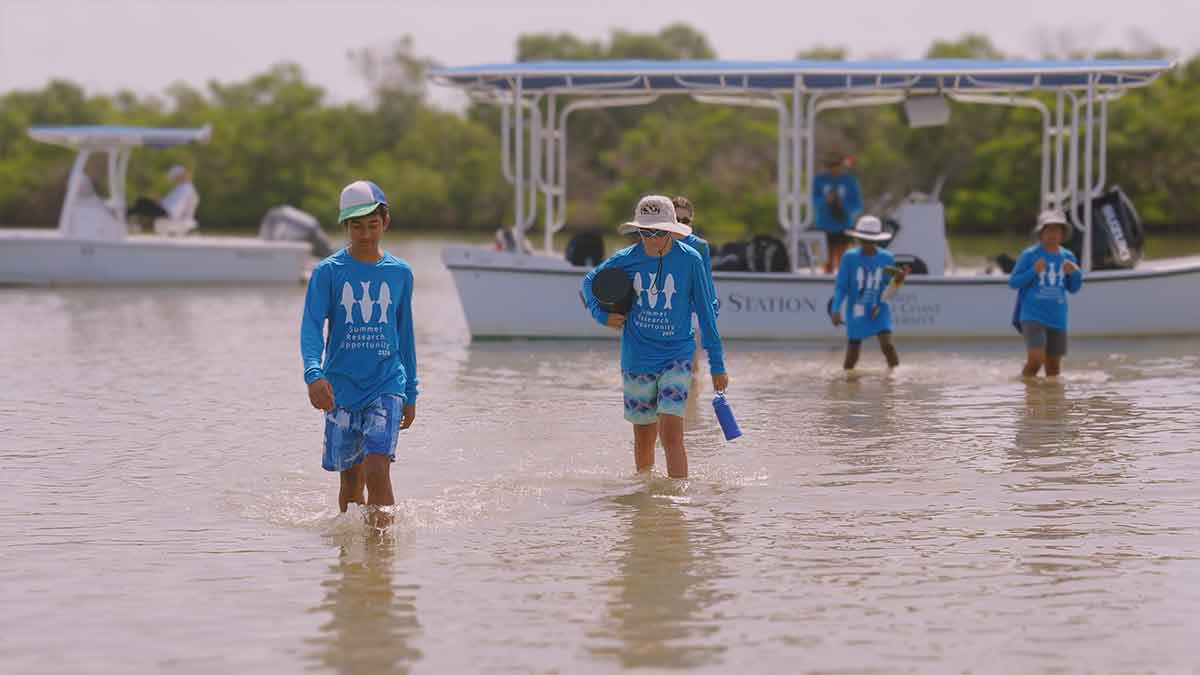 Several middle grade children in blue shirts are in knee-deep waters near a boat