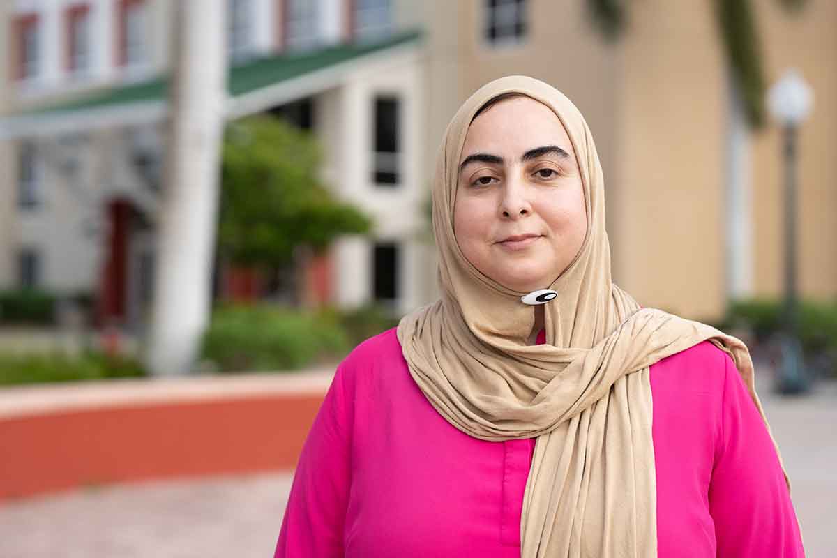 A woman in a pink top and beige head scarf stands outdoors in front of a residence hall