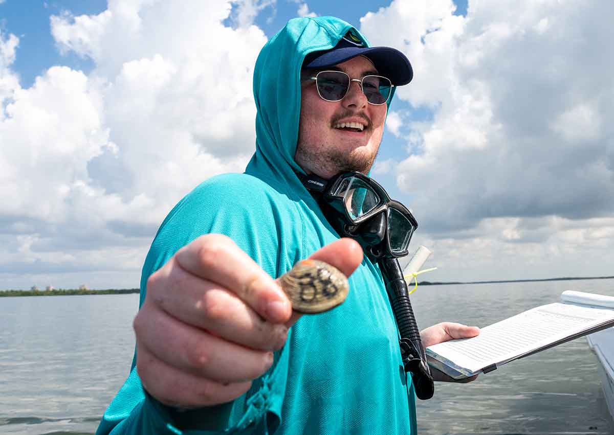 A young man in sunglasses, a ball cap and a teal hooded shirt holds a clam, with water and a cloudy sky in the background
