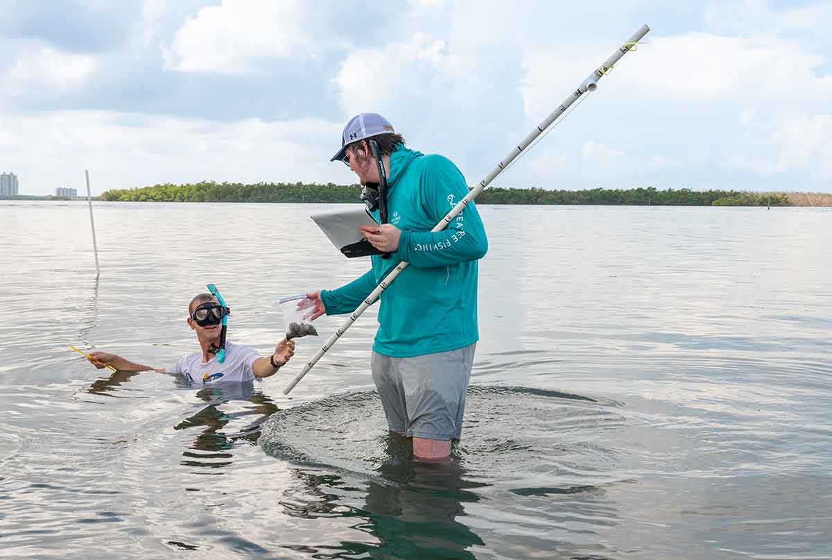 A man in snorkel equipment, submerged in water up to his chest, hands a clam to a young man in a teal hooded shirt stands