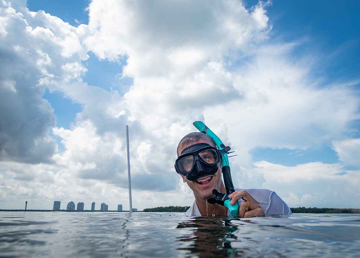 A man in snorkel equipment smiles. Only his head is out of the water, with a city skyline and large expanse of blue sky with clouds in the background