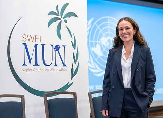 A woman in a navy blue blazer and slacks and white button-down shirt stands next to a sign that reads SWFL MUN Naples Council on World Affairs