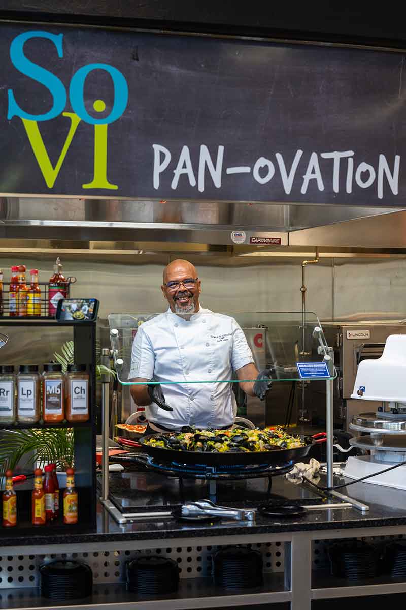A man in a white chef's jacket stands in front of an oversized paella pan under a sign that reads SoVi pan-ovation