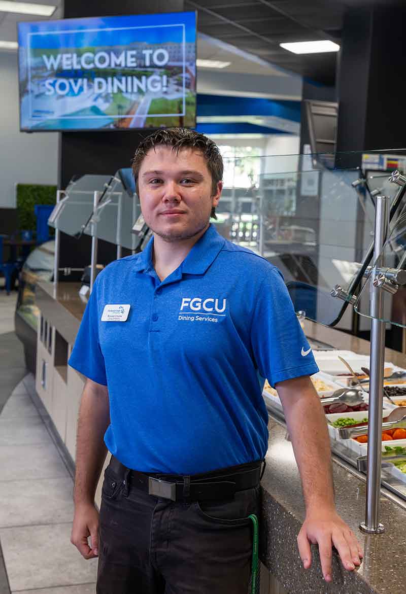 A young man in a blue polo shirt stands in front of a buffet in a dining hall. A TV behind him reads SoVi Dining and features an image of the FGCU campus