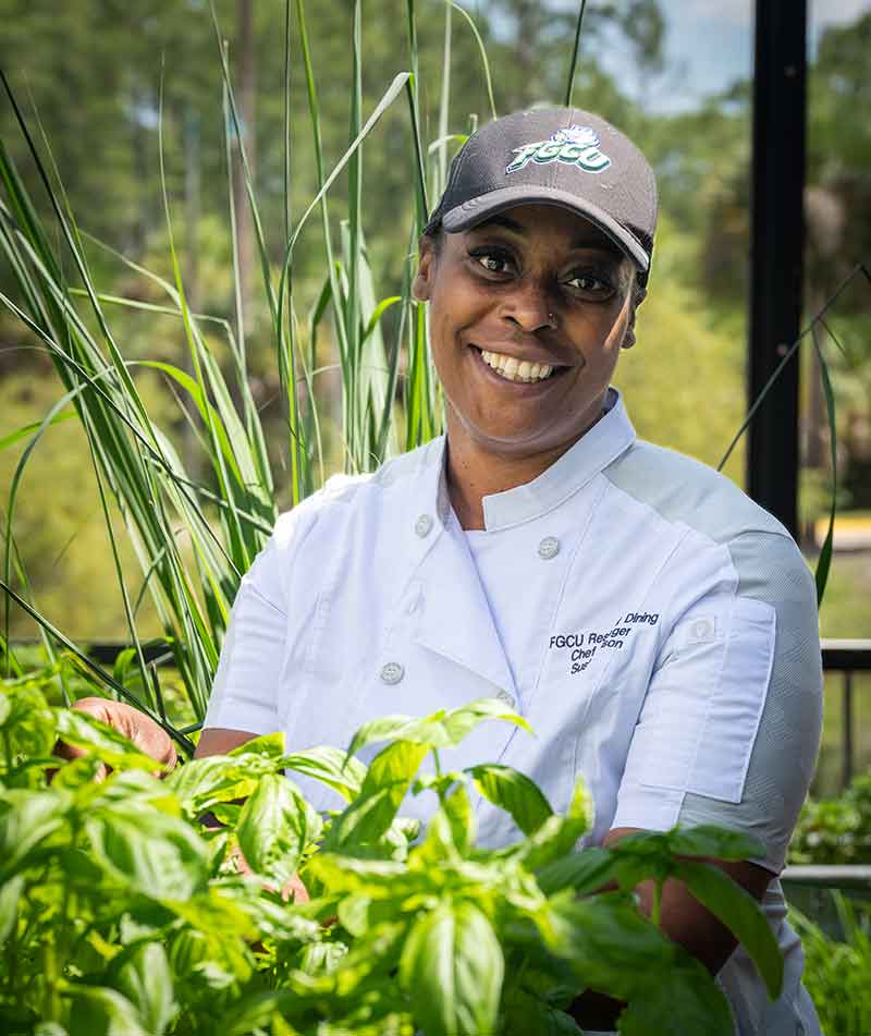 A woman in ball cap and white chef's jacket stands in a screened-in, outdoor area like a porch, holding uncut produce