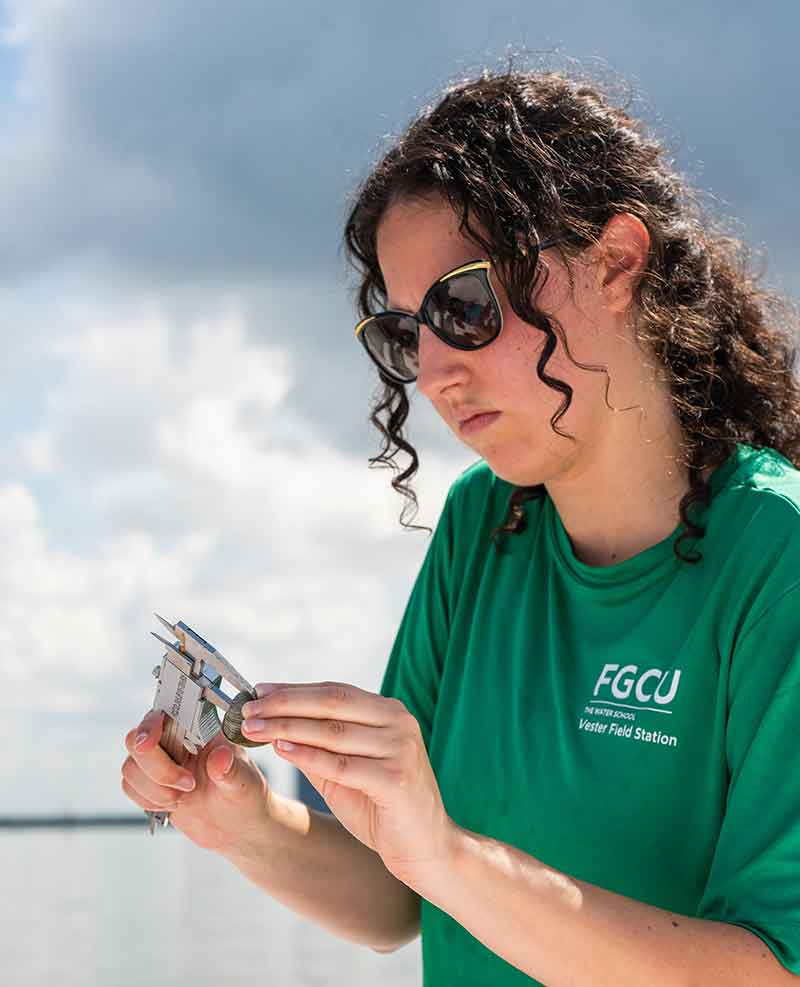 A young woman with curly hair, sunglasses and a green t-shirt holds a scientific measuring tool, with water and a cloudy sky in the background