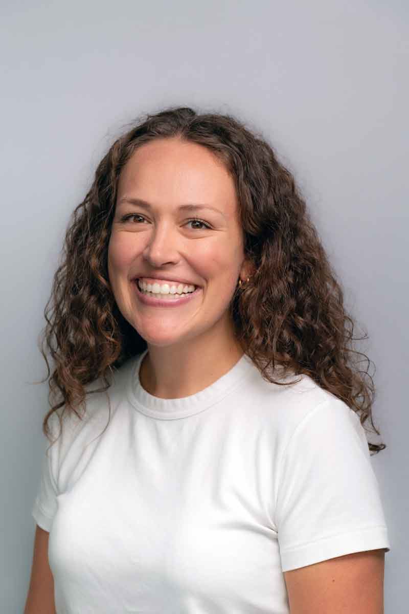 A headshot of a smiling woman with curly, brown hair in a white t-shirt