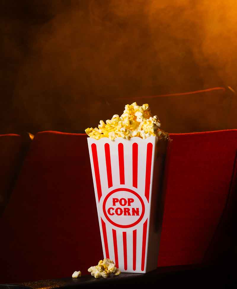 A slim red and white container has popcorn flowing up and over the rim, with a red, velvet cinema seat in the background