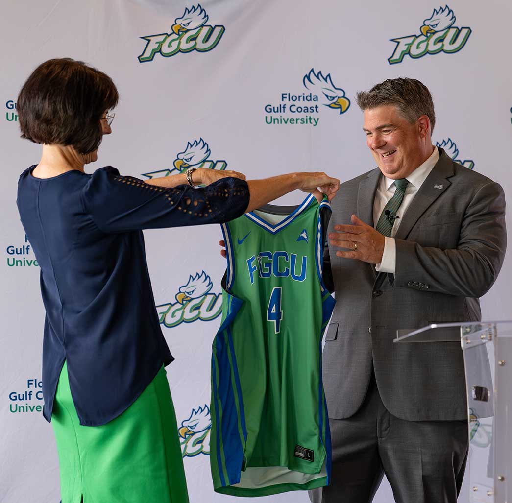 Person in blue top and green skirt presenting and FGCU jersey to a person in a gray suit