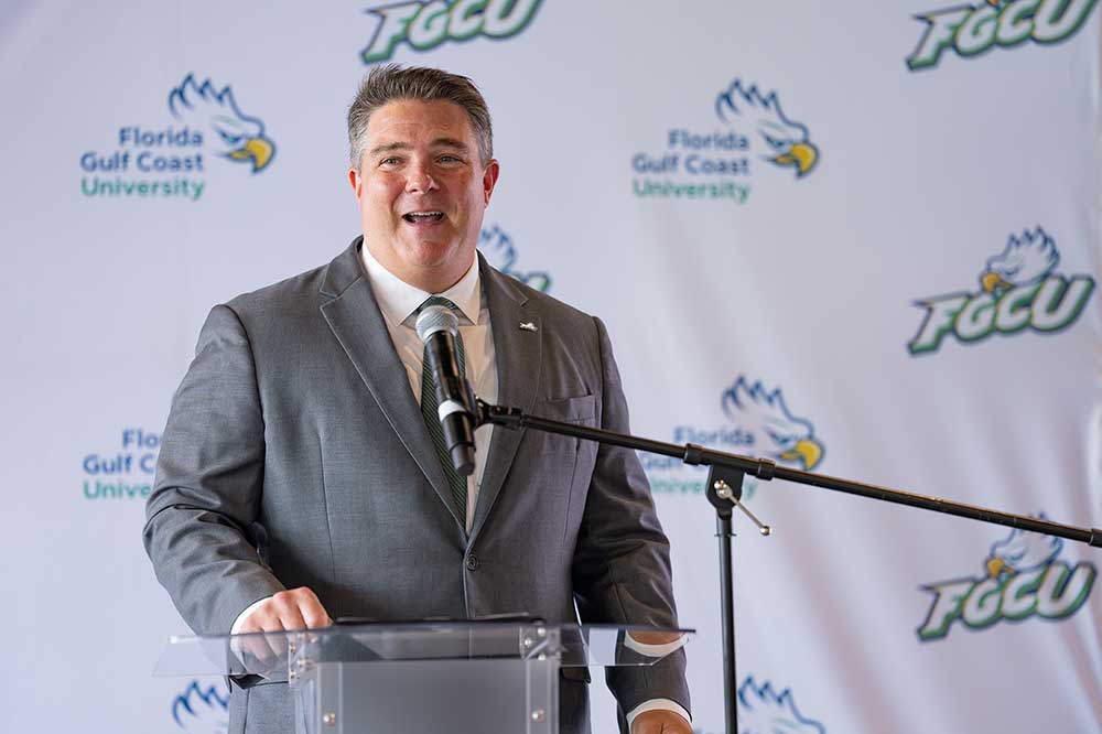 Person in gray suit speaking at a podium in front of and FGCU Athletics backdrop