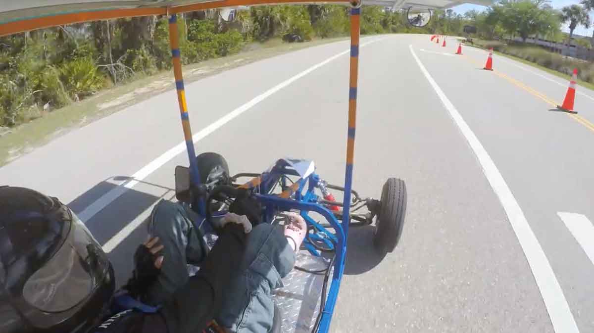 The view from a camera above the driver as a solar go-kart races around a road, with white lines on the pavement and orange safety cones lining one side