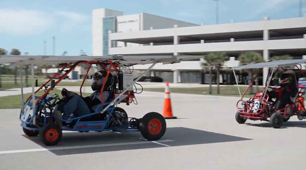 A solar go-kart with driver moving along a paved road in front of a large, grey parking garage on the FGCU campus