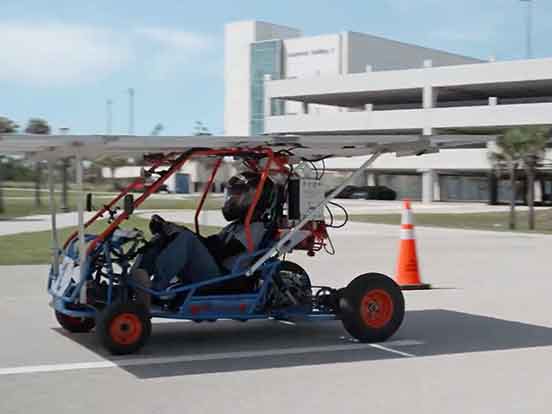 A solar go-kart with driver moving along a paved road in front of a large, grey parking garage on the FGCU campus