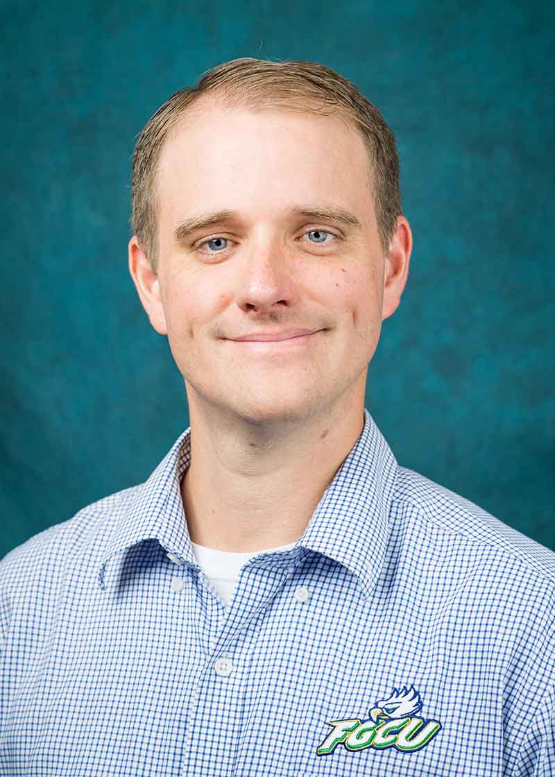 A headshot of a man in a light blue button-down shirt with the FGCU logo