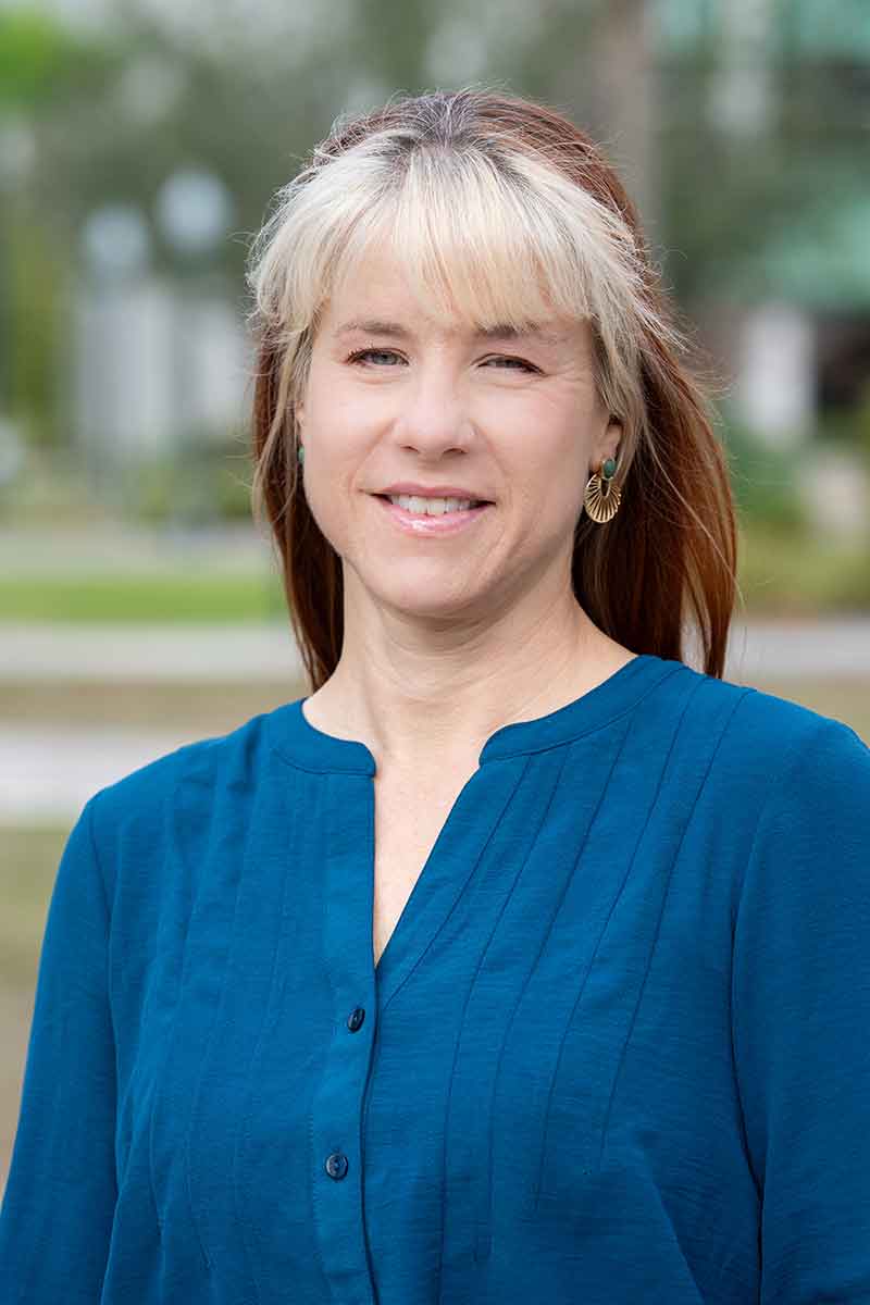 An outdoor headshot of a woman with pale blonde bangs and red hair in a blue shirt