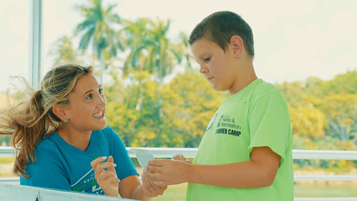 Outdoors, a smiling blonde woman in a blue shirt is looking up at a child in a green shirt