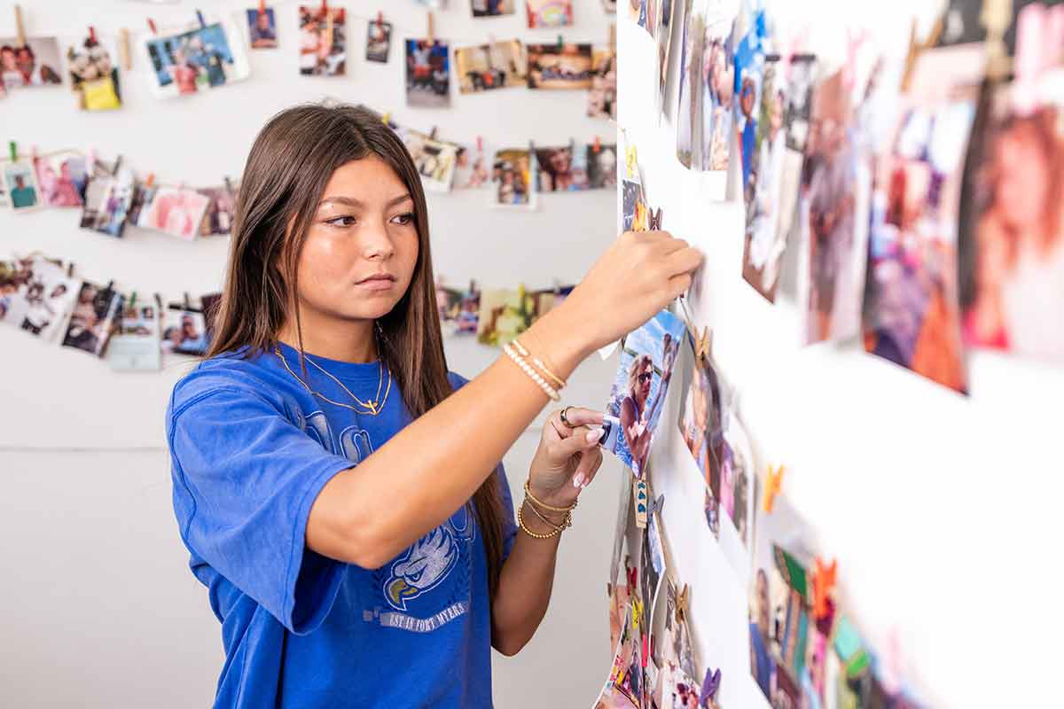 A young woman in a bright blue t-shirt hangs a photo on a wall covered in photos