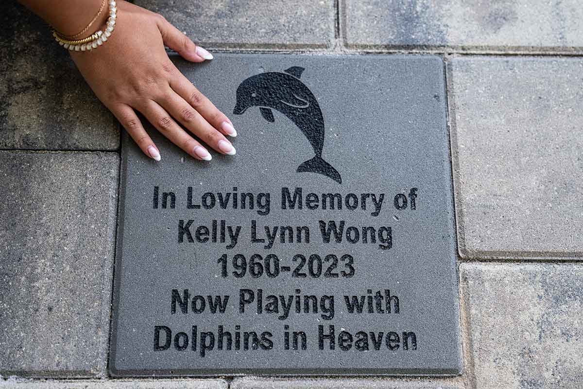 A photo of a hand over a paver that reads, In loving memory of Kelly Lynn Wong, 1960 - 2023, now playing with dolphins in heaven