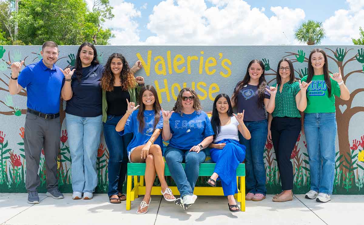 A group photo of nine people standing or seated on a bench outdoors, holding up their hands in the FGCU Wings Up gesture