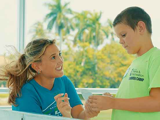 Outdoors, a smiling blonde woman in a blue shirt is looking up at a child in a green shirt