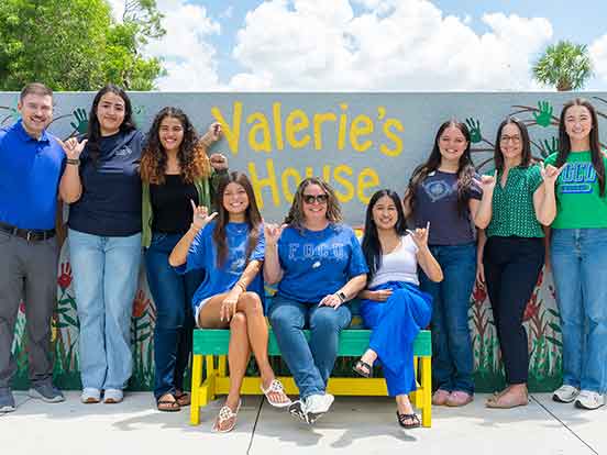 A group photo of nine people standing or seated on a bench outdoors, holding up their hands in the FGCU Wings Up gesture
