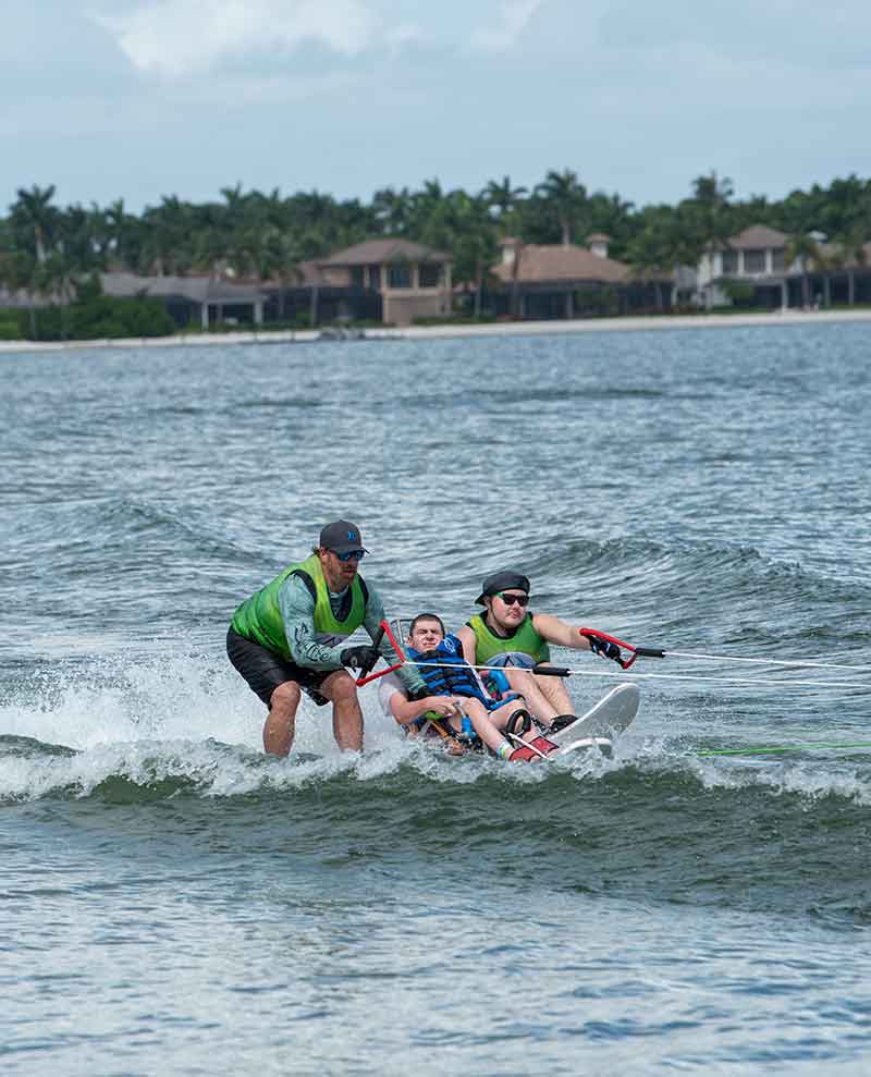 An outdoor photo in a large body of water: Two men in green tank tops and shorts assist a seated water-skier