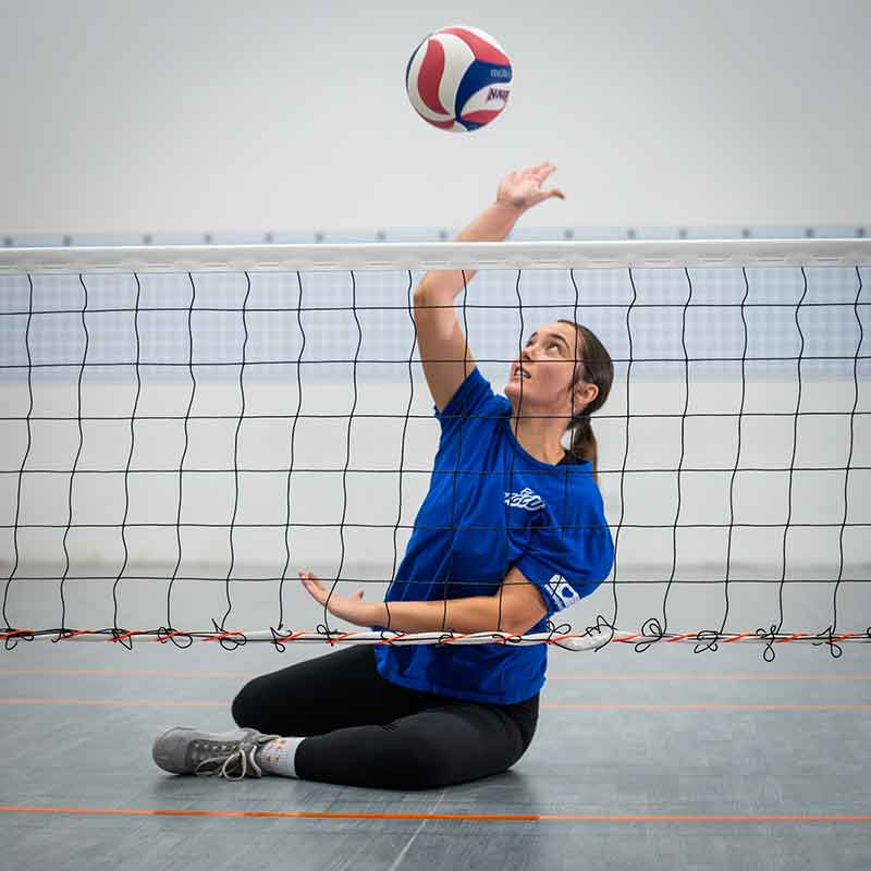 A young woman in a blue t-shirt and black yoga pants sits on the floor, hitting a red, white and blue volleyball over a net very low and near to the floor