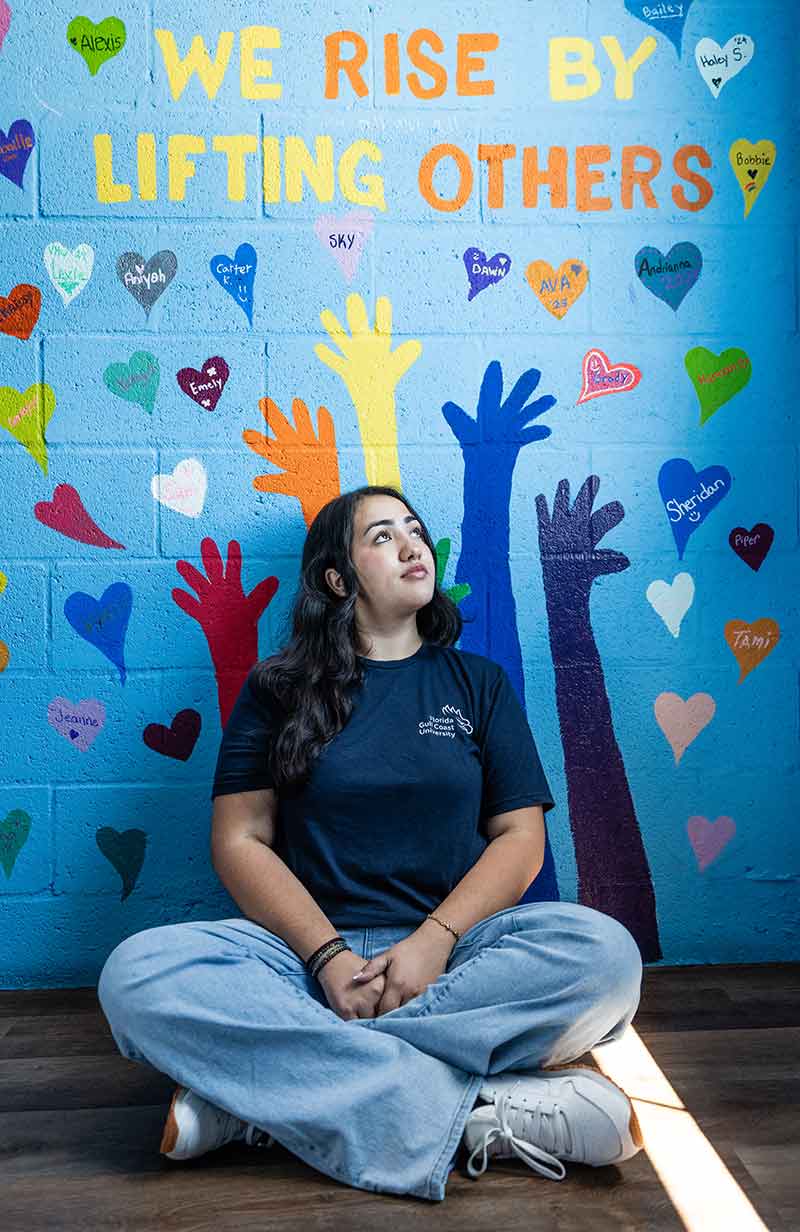 A young woman in blue jeans and a dark t-shirt sits on the floor in front of a wall with hands painted on the wall and a message that reads We rise by lifting others