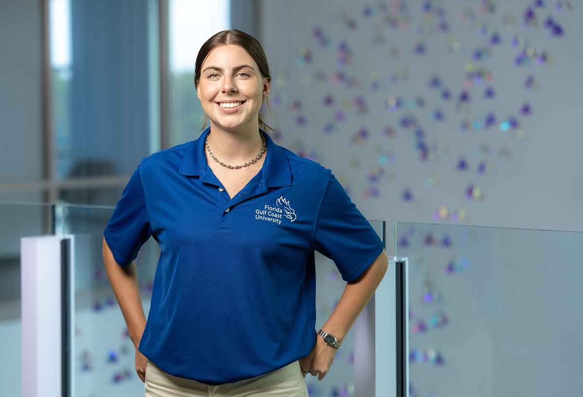 A smiling young woman in a blue FGCU polo shirt stands in front of a glass partition