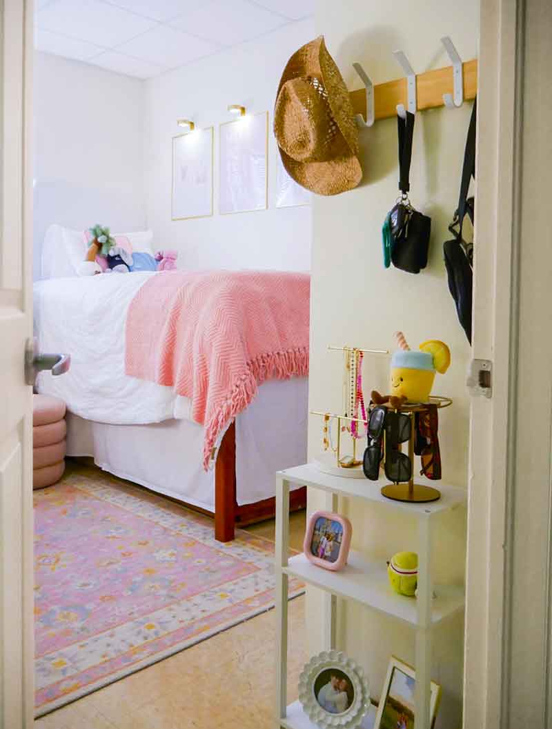 A bedroom decorated in white and pink palette, with a small nook in the foreground near the door and a raised bed with white and coral pink bedding in the background